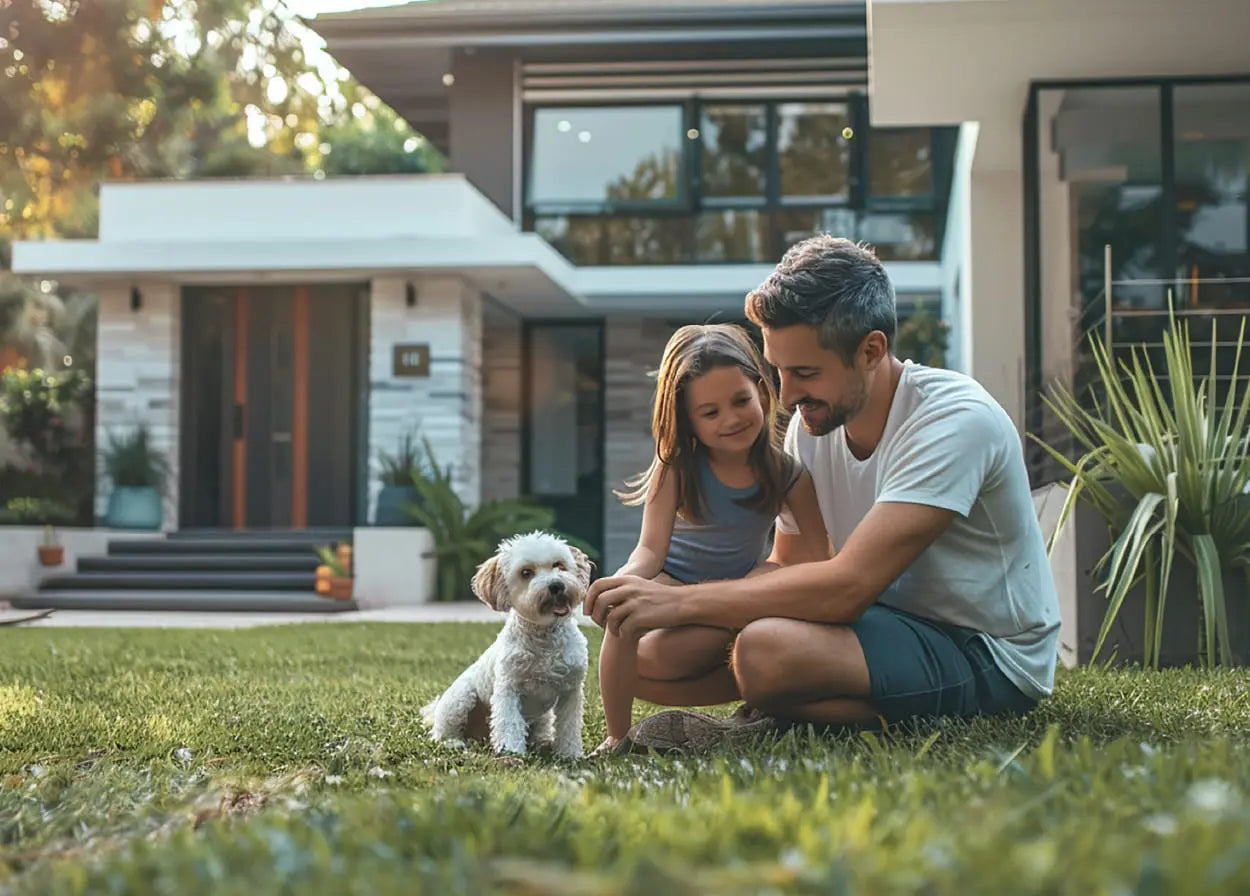 Man and young girl sitting on grass with a small dog in front of a modern house.